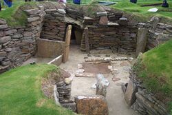 A jumble of stone walls, larger stones set into the ground and surrounding grassy areas. Various human figures wander past in the background.