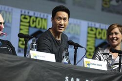 An Asian man with short black hair smiles towards the camera while sat behind a desk. A woman with short blonde hair sits next to him laughing. There are microphones on the desk in front of them.